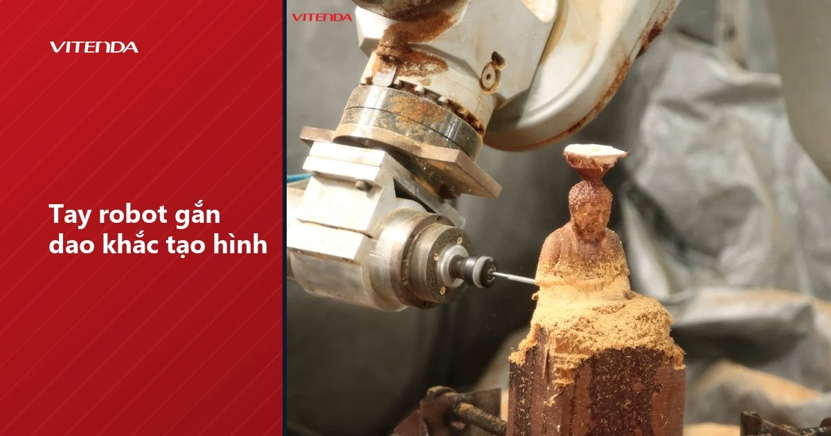 Cánh tay robot gắn dao khắc đang tạo hình một tượng nhỏ trên khối gỗ, mùn cưa bám quanh
